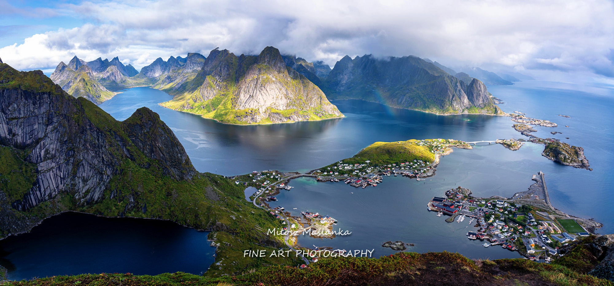 Sunny aerial panoramic view on stunning mountains and village of Reine in Lofoten islands, Norway from Reinebringen ridge. Scenic vista with a rainbow and rugged peaks.